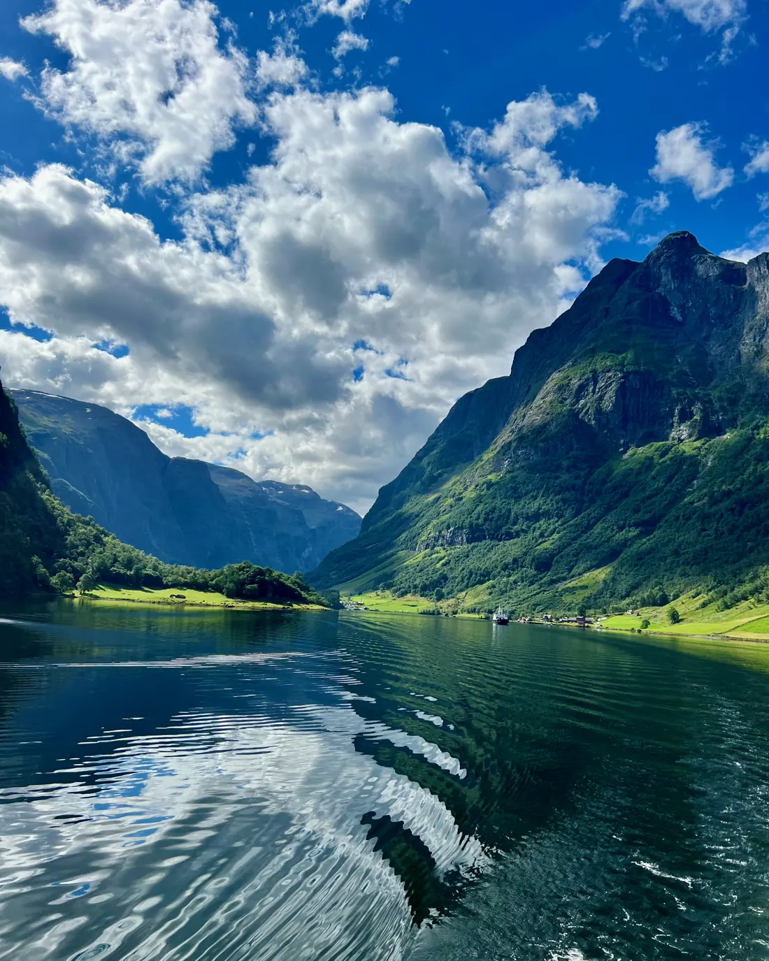 La estela de un barco en un fiordo verde, entre montañas altas y nubes.