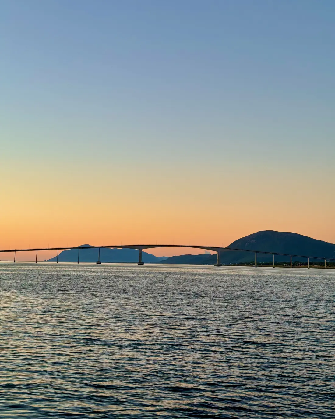 Un puente largo sobre el mar al atardecer, con el cielo naranja y una montaña al fondo.